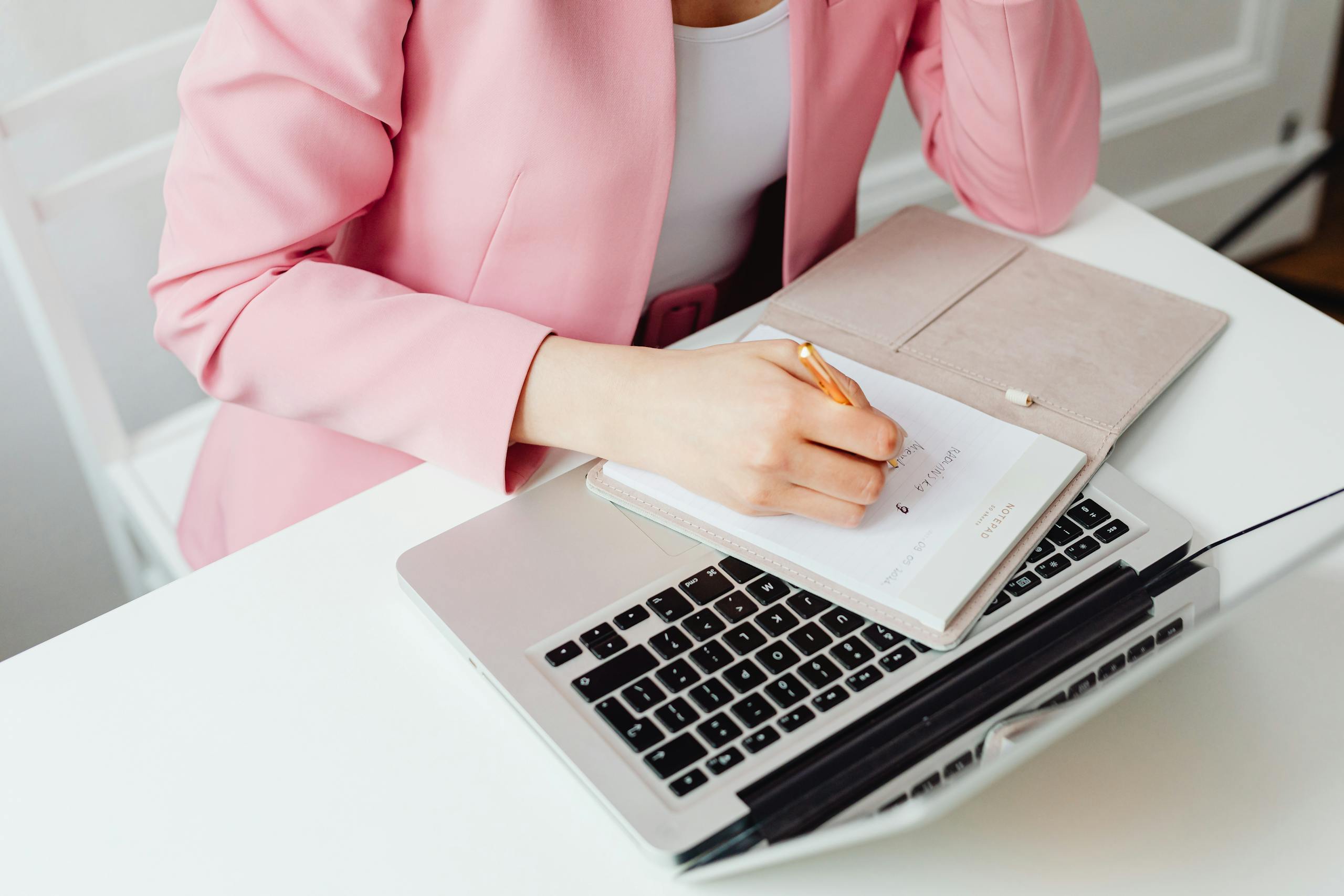 Professional woman in pink blazer writing notes on laptop for work efficiency.